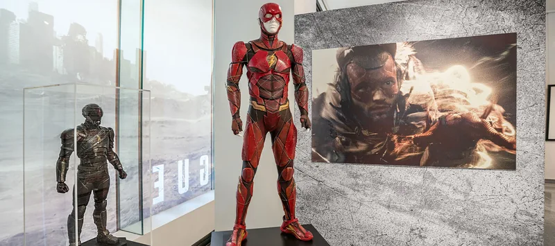 Traje de Flash en rojo junto a un traje oscuro y un panel con un fotograma de la película.